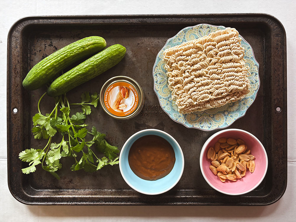 overhead view of ingredients including ramen noodles, mini cucumbers, peanut sauce, peanuts, cilantro and canned tuna